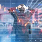 A dramatic conceptual image of a businessman viewed from behind, holding his head in his hands in a gesture of stress. He is standing in front of a large, semi-transparent digital overlay of a stock market dashboard. The overlay features a glowing orange and yellow candlestick chart showing a sharp downward trend, several fluctuating white line graphs, a world map, and percentage markers. The background shows a blurred office or trading floor environment.