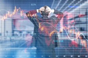 A dramatic conceptual image of a businessman viewed from behind, holding his head in his hands in a gesture of stress. He is standing in front of a large, semi-transparent digital overlay of a stock market dashboard. The overlay features a glowing orange and yellow candlestick chart showing a sharp downward trend, several fluctuating white line graphs, a world map, and percentage markers. The background shows a blurred office or trading floor environment.