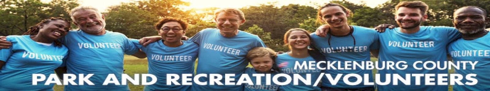 A diverse group of smiling people wearing bright blue volunteer t-shirts, standing with their arms around each other's shoulders with the sun shining through trees in the background. 