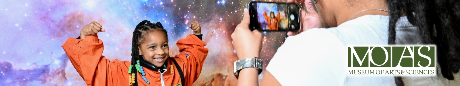 Child smiling inside a space suit getting her picture taken by her mother at the Museum of Arts & Sciences.