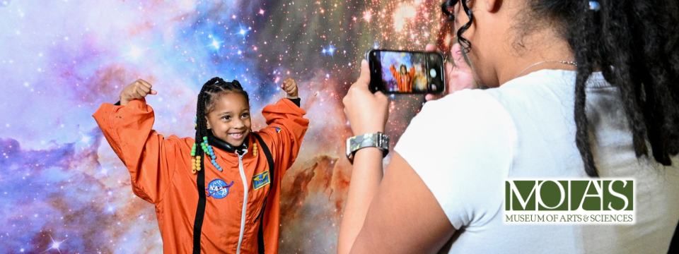 Child smiling inside a space suit getting her picture taken by her mother at the Museum of Arts & Sciences.