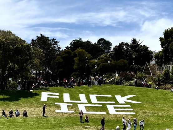 A large white-lettered message reading “FUCK ICE” is laid out across a grassy hill in a public park, likely a political protest or art installation opposing U.S. Immigration and Customs Enforcement (ICE). People are scattered across the lawn and gathered at the top of the hill, some taking photos or sitting in small groups. Trees and residential buildings are visible in the background under a partly cloudy sky.