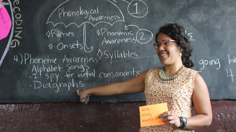 A Puerto Rican woman stands teaching at a blackboard, smiling.