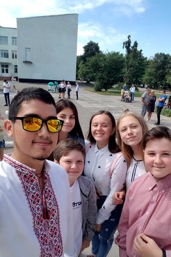 A young Mexican American man wearing traditional Ukrainian clothes stands smiling with his young, Ukrainian students outside.