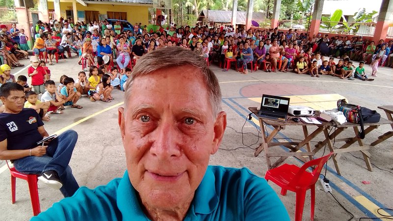 An older American white male takes a selfie in front of a large group of his primary school students and fellow teachers.