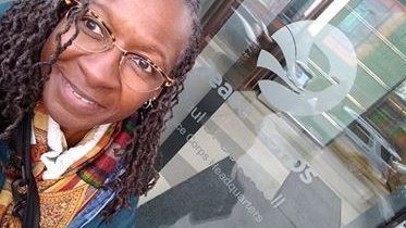 An older Black woman smiles for a selfie in front of a Peace Corps logo on a window.