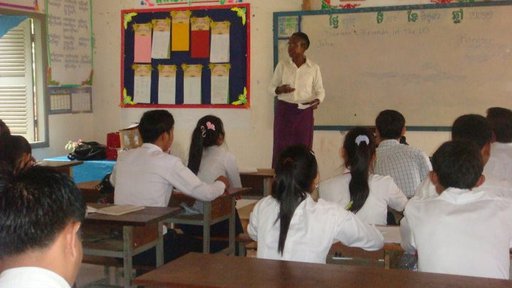 An older, Black woman stands in front of a classroom teaching Cambodian students.