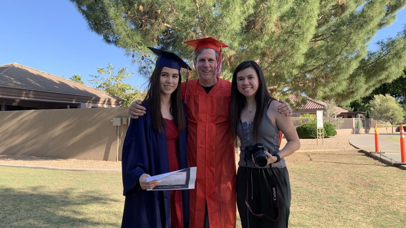 Three people stand together outside. The woman on the left and the man in the middle are wearing graduation gowns