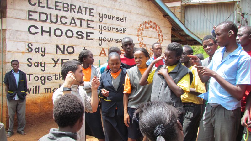Male Peace Corps Volunteer stands outside a school, signing to deaf students.