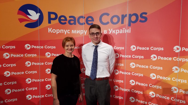 A man and a woman smile next to each other in front of a large orange Peace Corps banner.