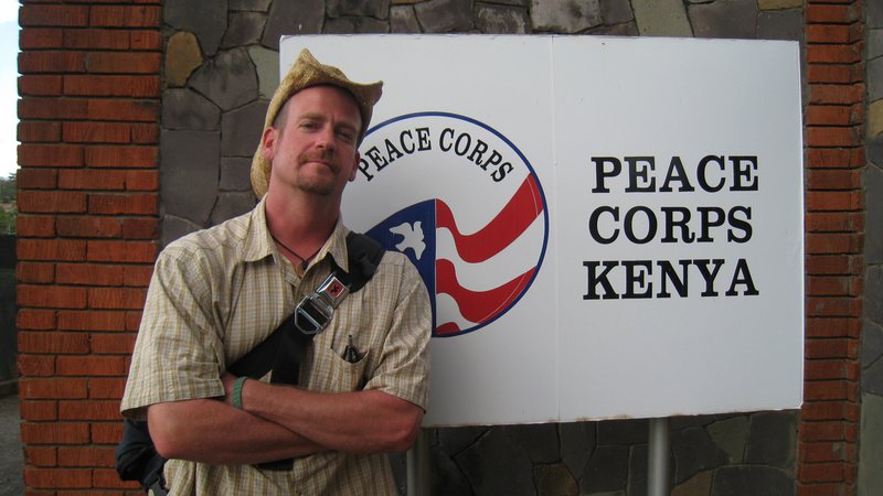 A young, white man wearing a cowboy hat stands outside in front of a Peace Corps Kenya sign.