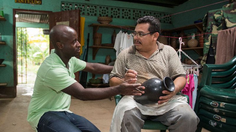 A man kneels next to another man seated on a chair inside.