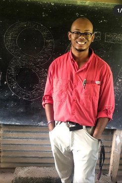 A Black American man in dress clothes stands in front of a chalkboard