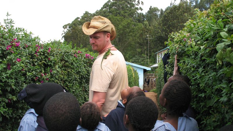 A young, white man shows off a chameleon on his shoulder to a group of young Kenyan students
