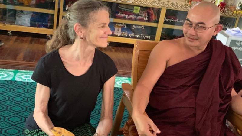 An older white women kneels at the feet of a monk sitting in a chair, offering him bananas.