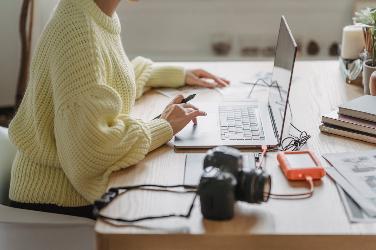 A person working on a laptop at a desk with a camera and hard drive nearby, representing digital publishing and content creation.