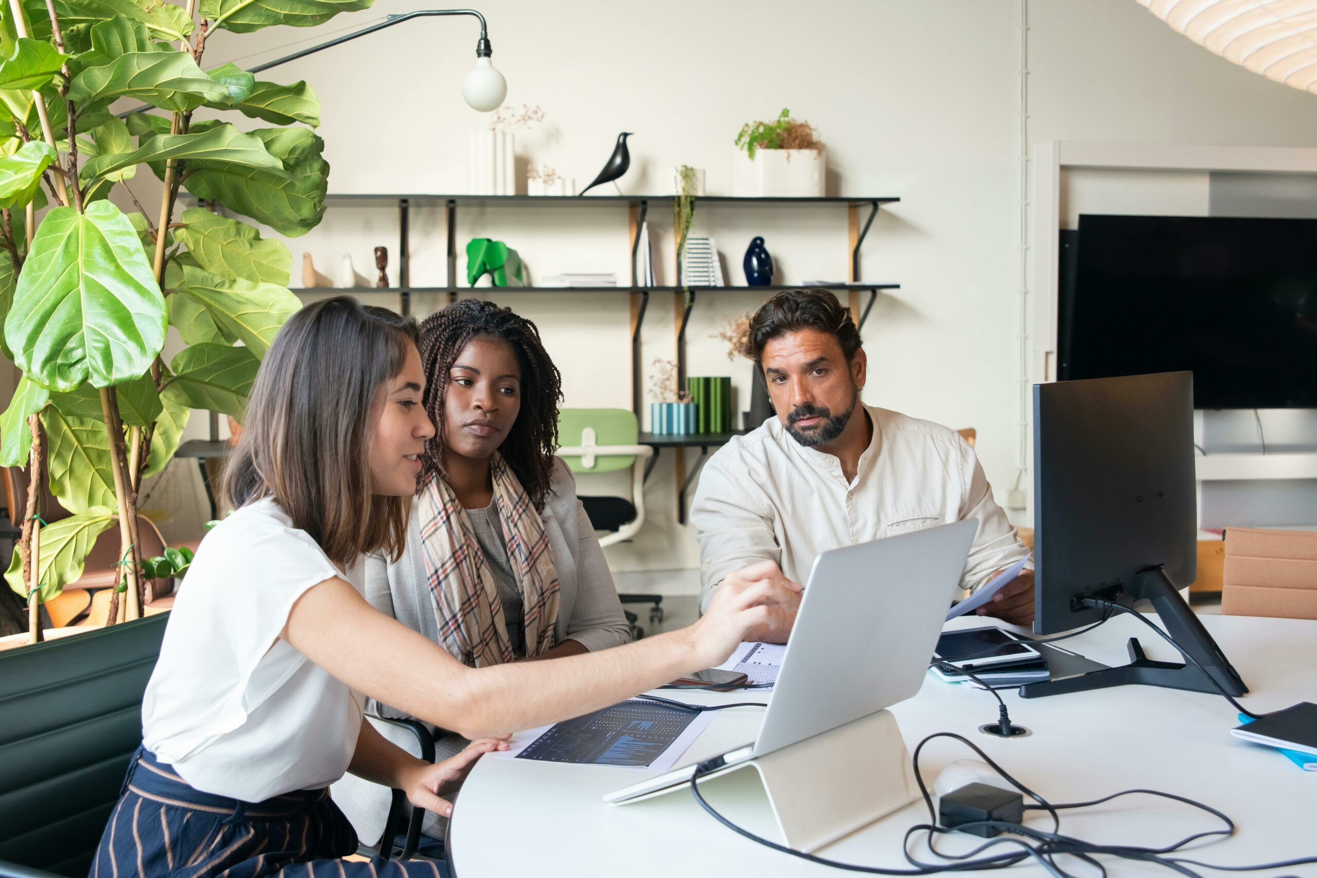 Three colleagues collaborating in a modern office, reviewing data and strategy on laptops and monitors surrounded by natural light and plants.