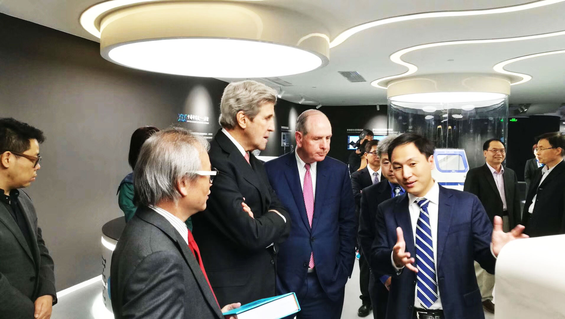 Él Jiankui explica el secuenciador del genoma construido por una de sus compañías, Direct Genomics, al ex secretario de estado estadounidense John Kerry.