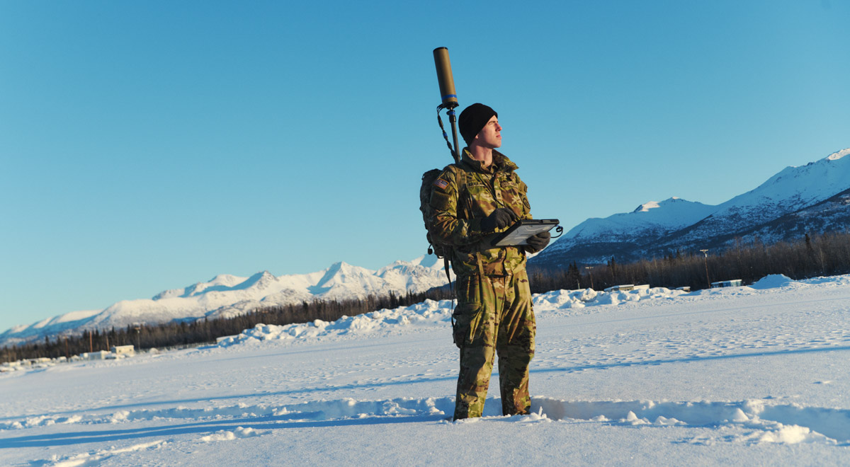 A service member stands in a snowy landscape holding a tablet, with a communications device on his back.