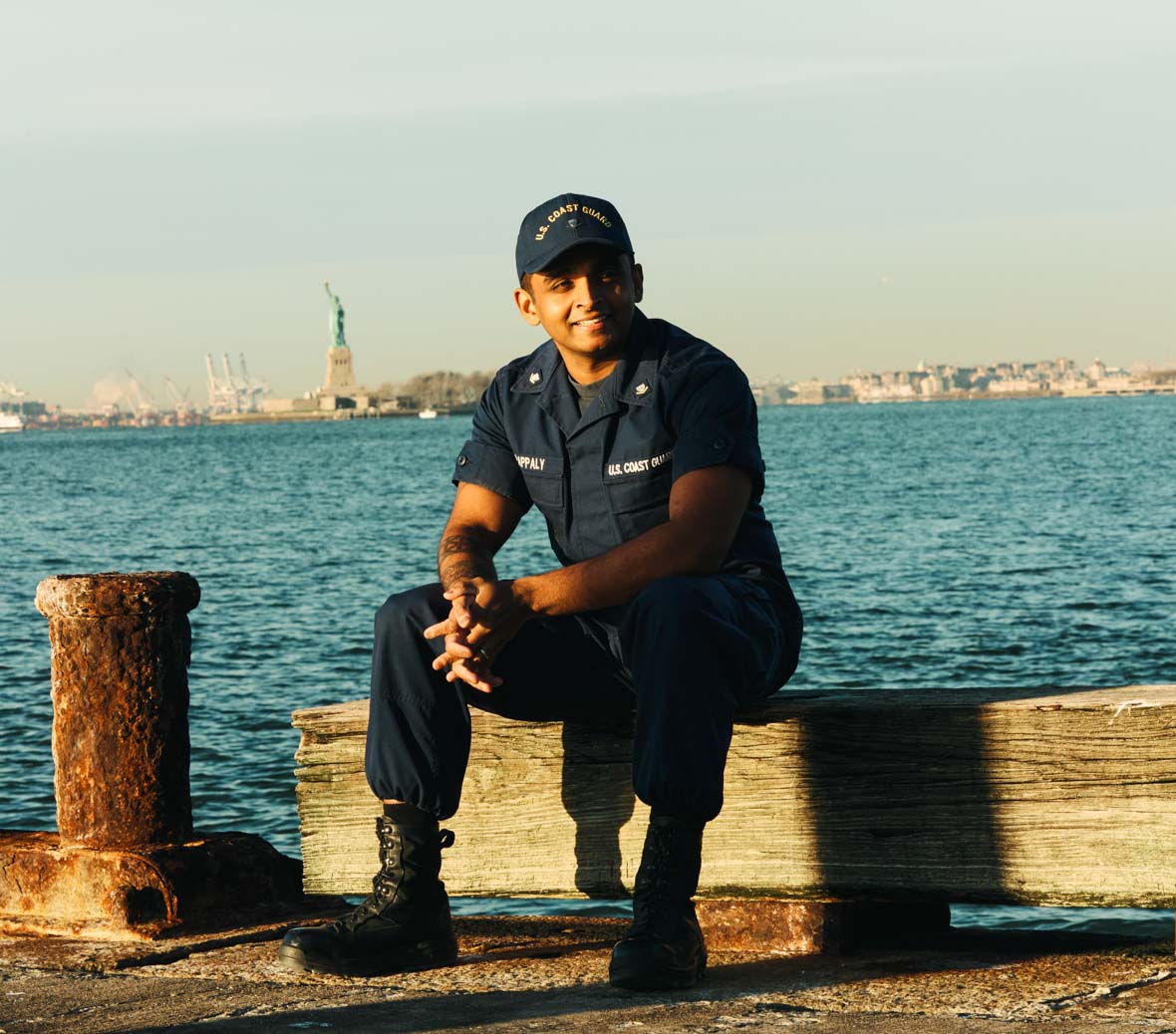 anthony sits by the water in NY with the statue of liberty in the background