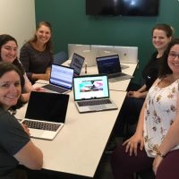 Five women seated around a table with laptops opened