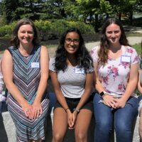 Five women wearing name tags and seated on a bench with trees in background