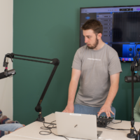 Student assists with the Rodecaster sound board while two students wearing headphones test their microphones