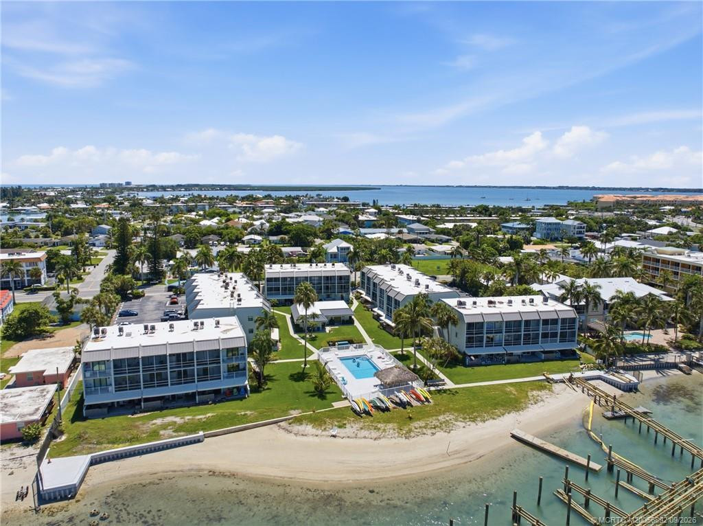 Aerial view showcasing the community’s waterfront setting, pool, sandy shoreline, and surrounding coastal neighborhood.