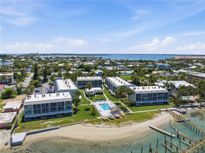 Aerial view showcasing the community’s waterfront setting, pool, sandy shoreline, and surrounding coastal neighborhood.