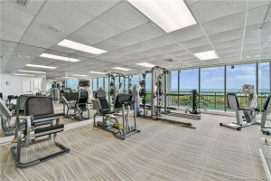 Well-equipped oceanfront fitness center featuring exercise machines, mirrored walls, and expansive windows overlooking the beach.