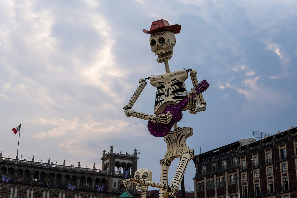 A towering Day of the Dead skeleton sculpture holding a purple guitar rises over Mexico City’s historic center, surrounded by colonial architecture and the Mexican flag. This vibrant cultural scene reflects the setting of the Fluenz Spanish Immersion in Mexico City, recognized as the best Spanish immersion experience for adults seeking to master the language through culture and tradition.