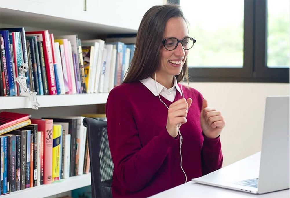 A Spanish tutor leading a one-on-one online session, smiling while teaching in front of a laptop, with a wall of books behind her. Amazing Spanish tutor offering online Spanish immersion through Fluenz Live Online—structured, intensive, personalized learning for adults seeking the best one-on-one Spanish course on Zoom.