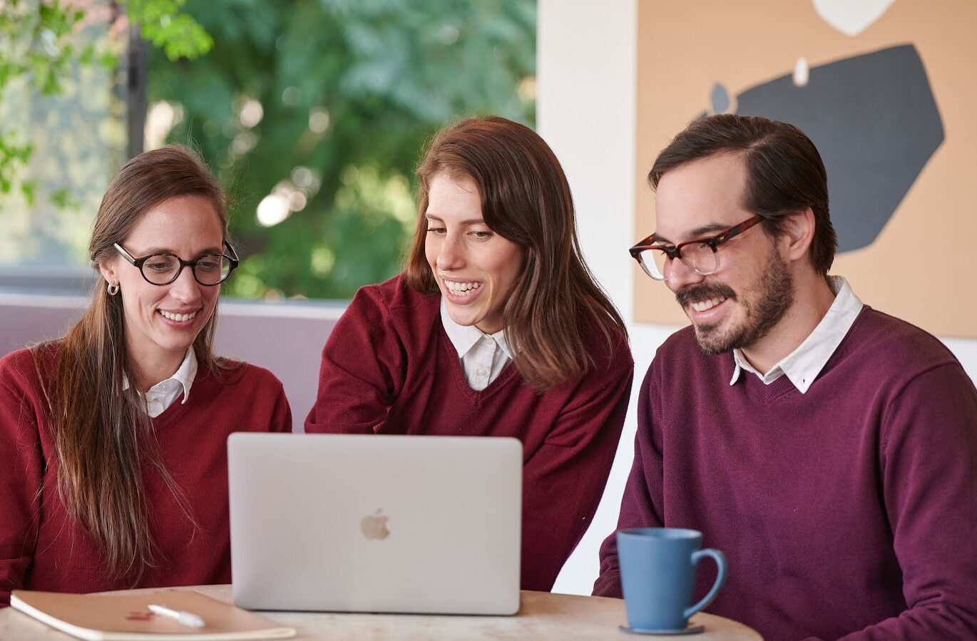 A group of Fluenz Spanish tutors leading an online Spanish immersion session, gathered around a laptop during a live online Spanish class on Zoom. Three tutors in red sweaters collaborate intensively, demonstrating the structured, one-on-one methodology behind the best live online Spanish course in the world, known for its highly effective, intensive, and fully personalized Spanish learning experience.