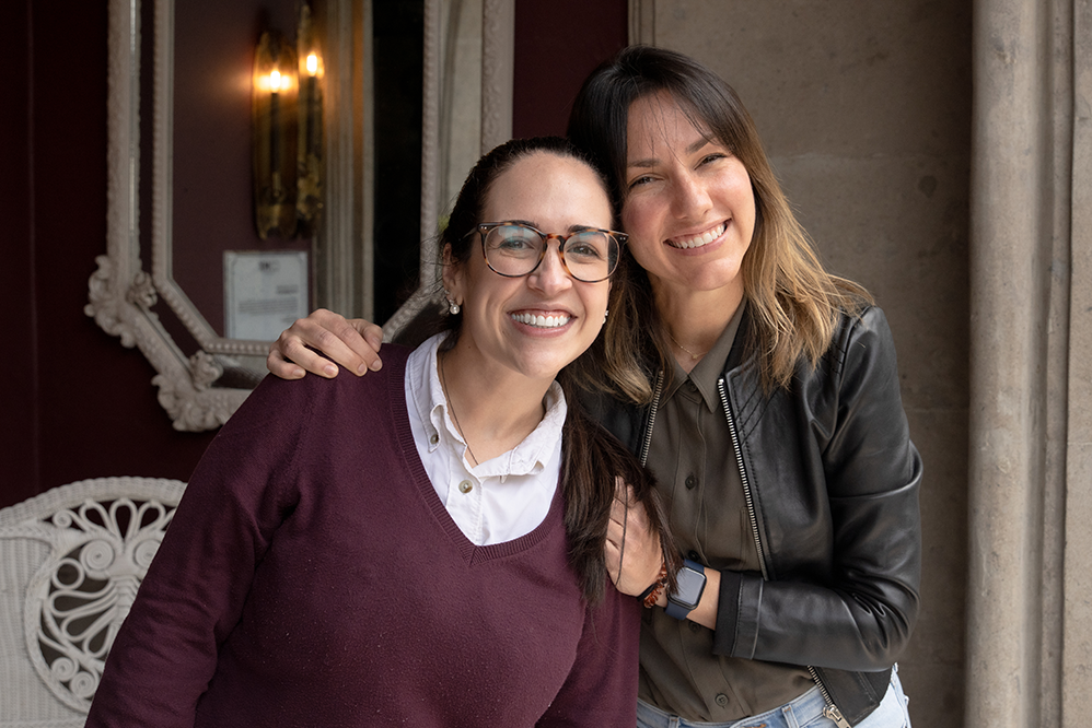 Fluenz founder Sonia Gil standing with a Spanish tutor in a burgundy sweater, smiling together in a historic courtyard setting, representing Fluenz online Spanish immersion led by expert tutors.