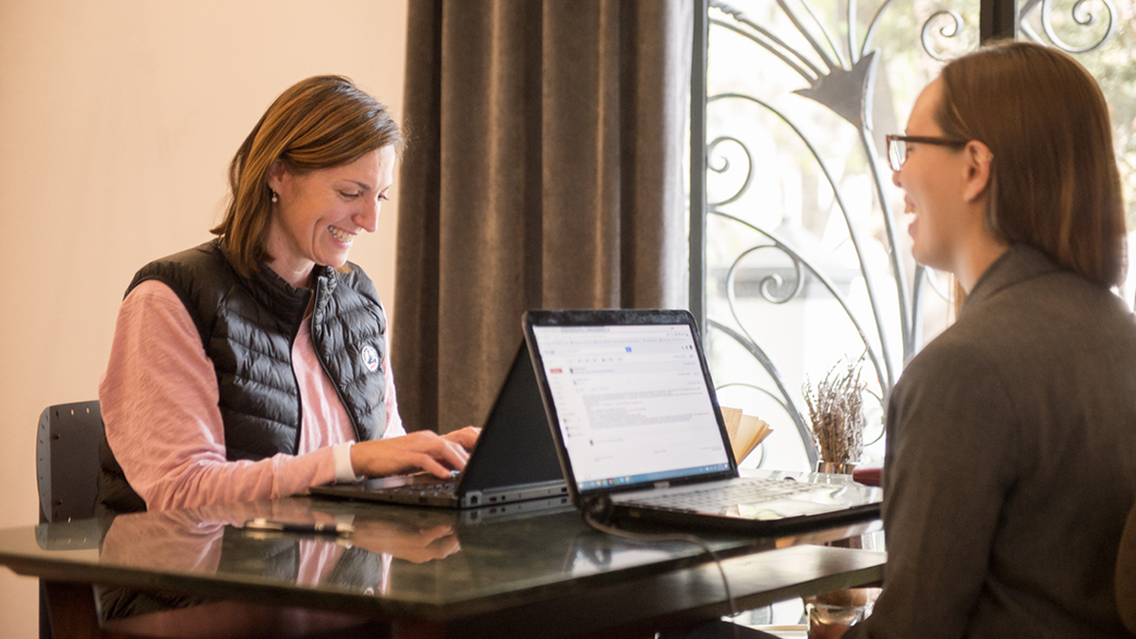 Two women engaged in a one-on-one Fluenz Spanish Bootcamp session, smiling and working on laptops at a glass table in a bright, elegant learning space.