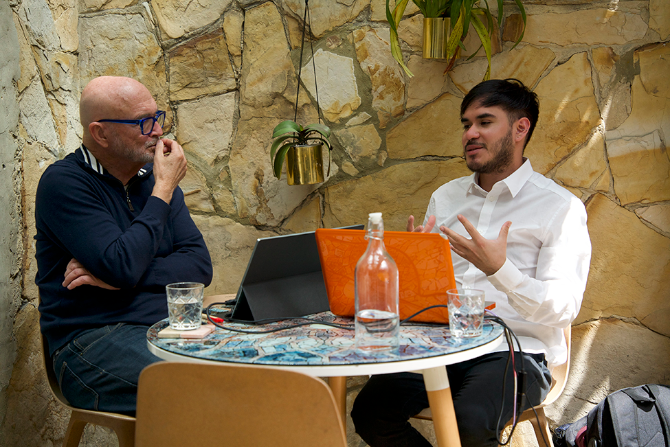 Adult learner and Spanish tutor engaged in a one-on-one Fluenz Spanish Bootcamp session, seated at a small table with laptops in a stone-walled courtyard, having a focused conversation during personalized instruction.