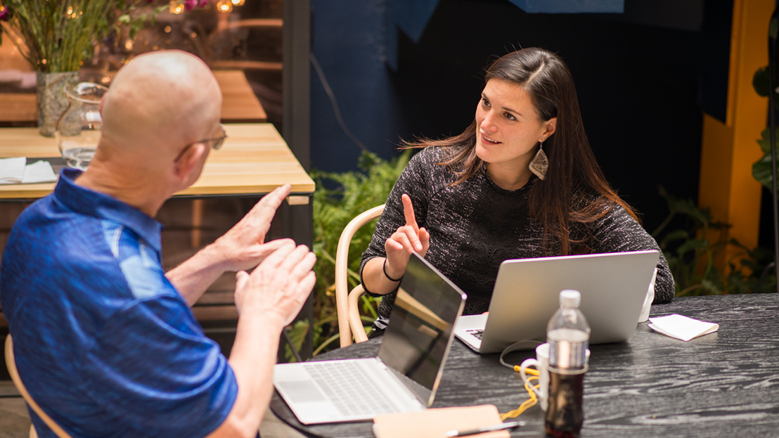 Spanish tutor guiding an adult learner during a one on one Fluenz Spanish Bootcamp session, both seated at a table with laptops in an outdoor setting.