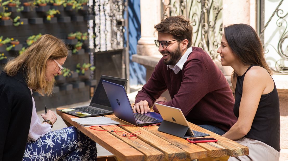 A Fluenz Spanish Immersion session in Mexico City unfolds at an outdoor wooden table, where an adult learner reviews notes while two tutors smile and collaborate using laptops and a tablet.