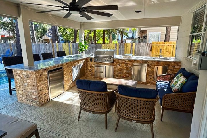 Covered outdoor kitchen with natural stone base, polished stone countertop, stainless grill, and nearby pool seating