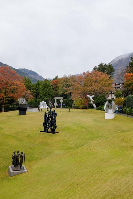 A green hill with modern art sculptures on the lawn and fall foliage in the background