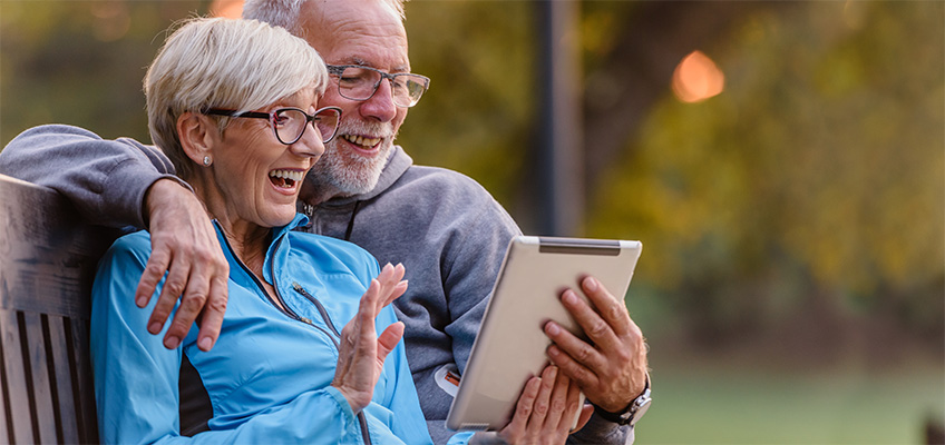 Two seniors sitting on a park bench holding a tablet in front of them.