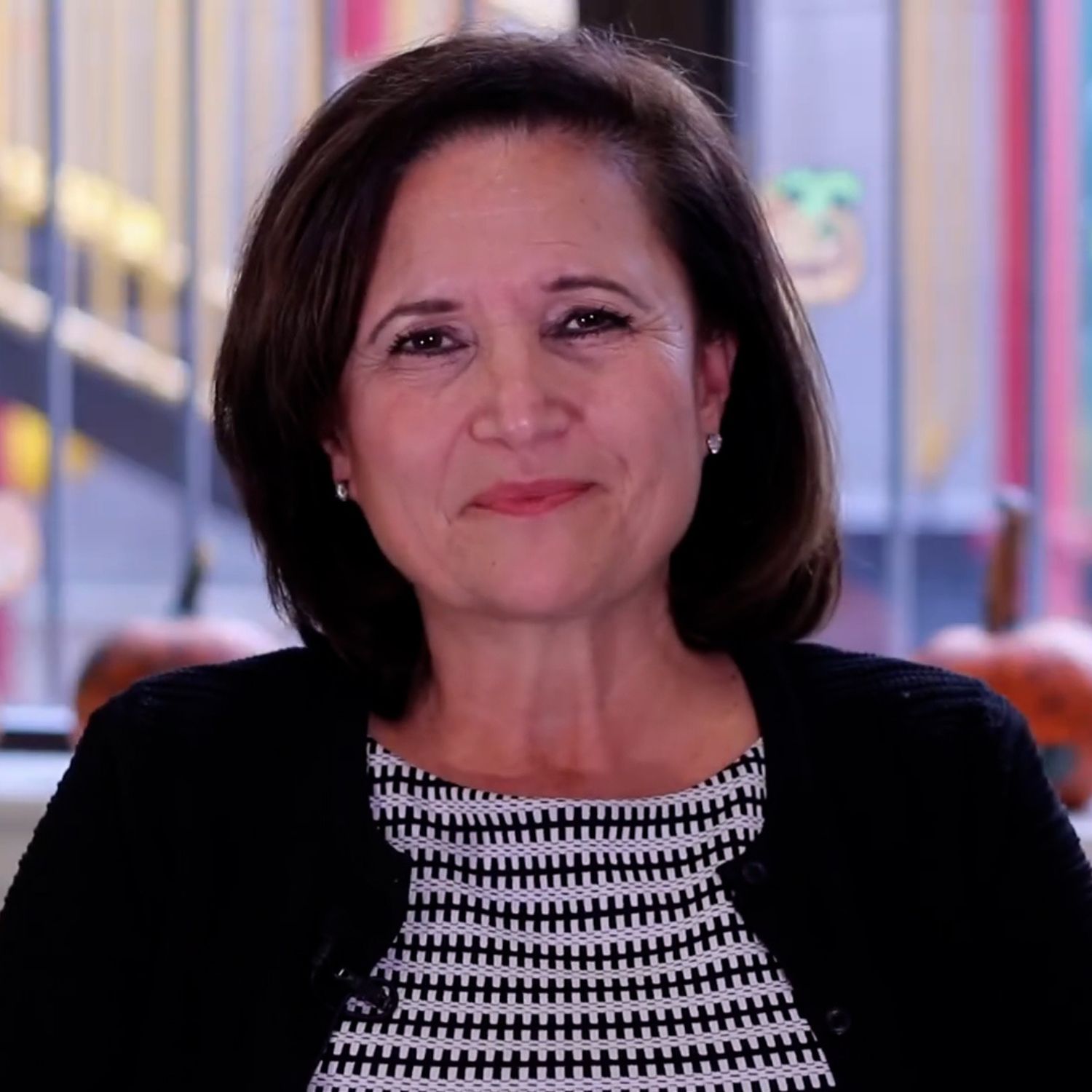 Portrait of a person with short, dark hair wearing a black jacket over a patterned top, seated indoors. The background is softly blurred with colorful elements and window light behind them.