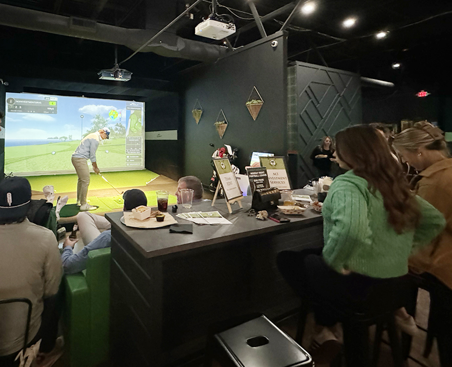 Group sitting at a table facing a golf simulator with someone swinging.