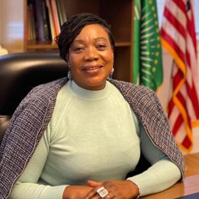 Amb. Suka-Mafudze is wearing a green top with grey shawl as she sits at a desk.