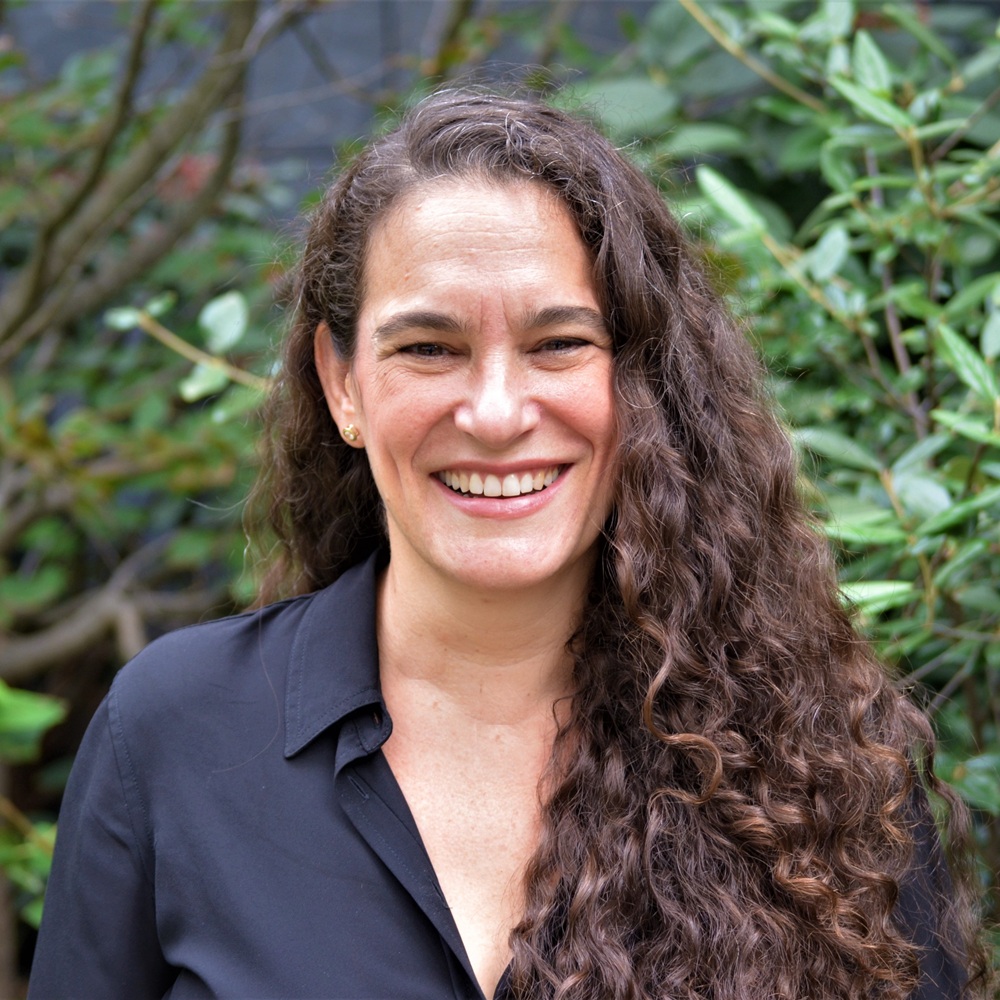 Portrait of an adult person with long, dark, curly hair wearing a black collared shirt, standing outdoors in front of green leafy plants.