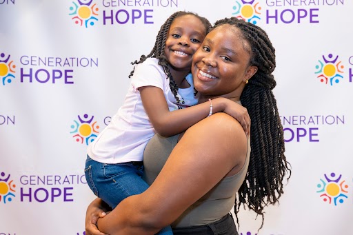 Generation Hope Scholar holding her child while both smile at the camera in front of a Generation Hope logo backdrop