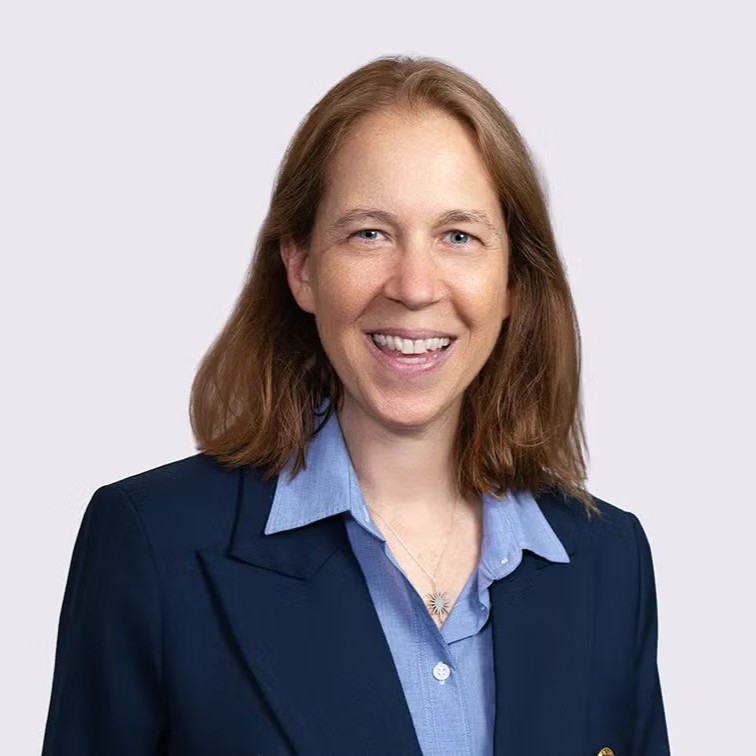 Head-and-shoulders studio portrait of an adult with shoulder-length hair, wearing a navy blazer over a light blue collared shirt, against a light neutral background.
