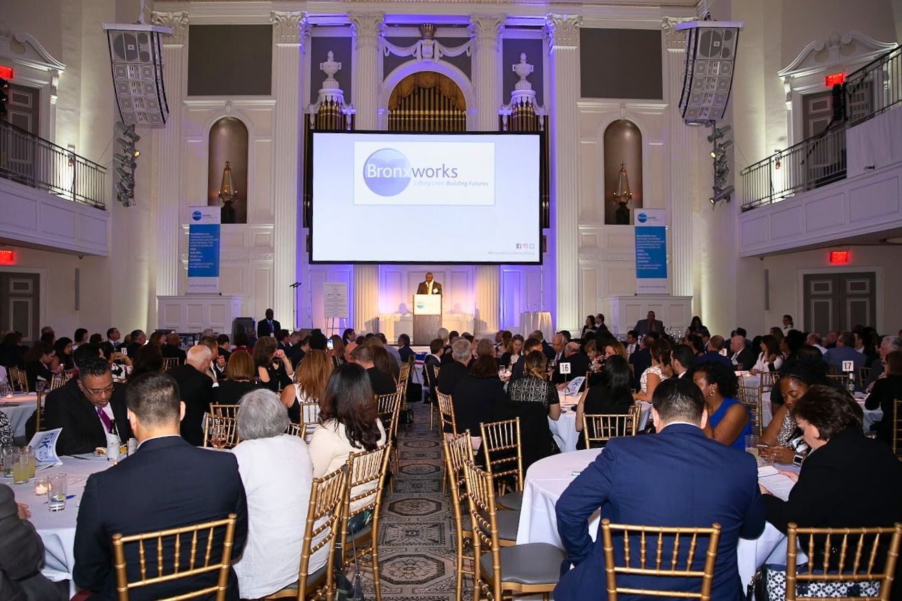 A large formal event in an elegant ballroom, with many people seated at round tables facing a stage. A speaker stands at a podium beneath a large screen displaying 