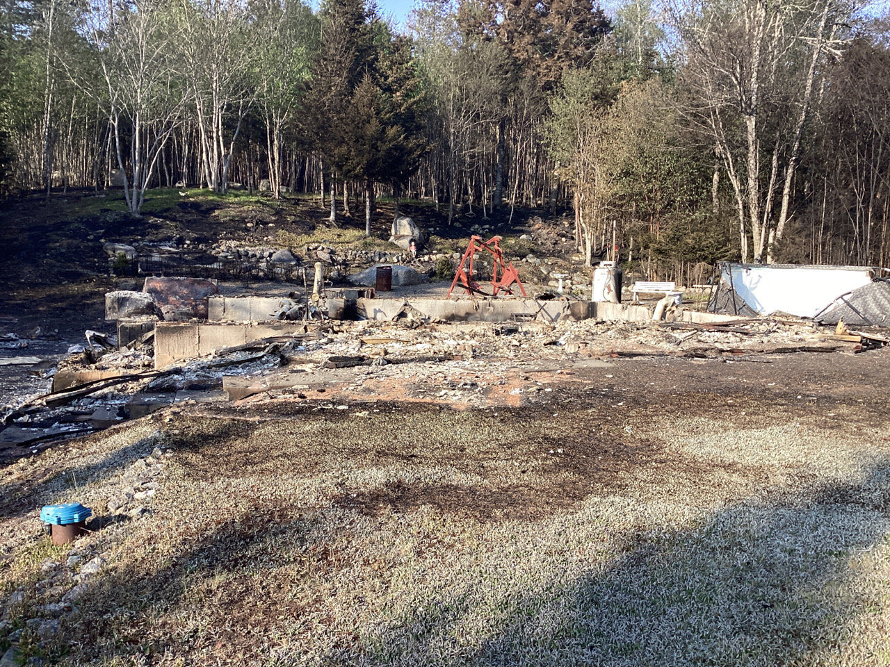 Phil and Sue's property after the fire: The remains of a house are shown in ruins, with charred debris and partially standing walls. The surrounding area is scorched, with burnt trees and vegetation, reflecting the aftermath of the Upper Tantallon wildfire.