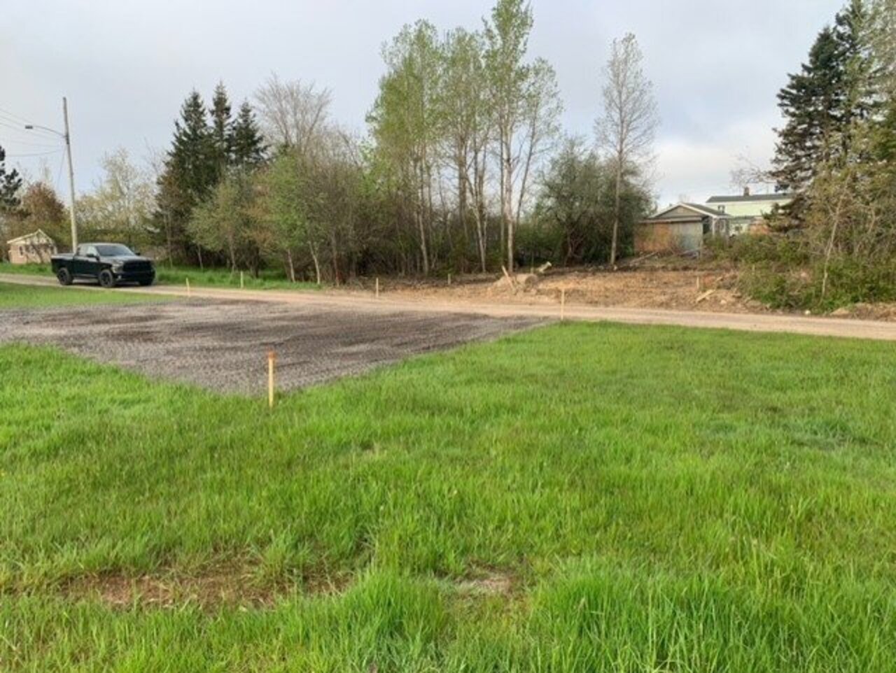 A prepared plot of land in New Glasgow, designated for a new Habitat for Humanity build. The ground is leveled and marked with stakes, ready for construction. Surrounding the plot are green grass, trees, and a nearby road with a black truck parked on the side.